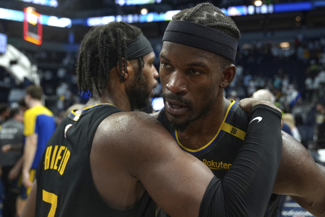Two basketball players in black jerseys embrace on the court. One has his back to the camera, while the other looks focused. The background shows a cheering crowd.